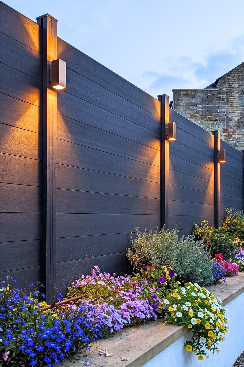 21. Black Composite Privacy Fence Design With Warm Wall Lights and Colorful Raised Flower Bed